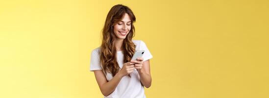 Cheerful lovely girlfriend texting friend pleased cute smile tap smartphone screen smiling broadly look mobile phone display tenderly writing post contemplate touching photo yellow background