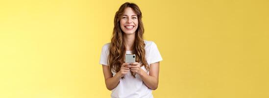 Cheerful carefree happy lively girl hold smartphone using awesome funny app playing cool game relaxing waiting friend park browsing social media using mobile phone look camera upbeat smiling photo