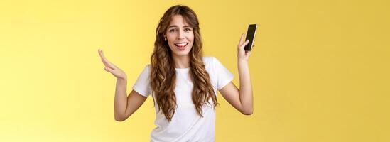 Carefree good-looking lively sociable curly-haired girl having fun wearing wireless earbuds dancing joyfully listening music moving rhythm song hold smartphone raising hands up smiling camera amused photo