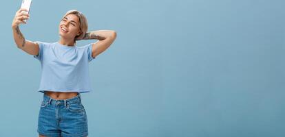 New post in my blog. Stylish feminine and sociable good-looking young female posing for selfie with pulled arm and smartphone sticking out tongue while making faces at device screen over blue wall photo