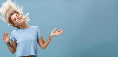 Waist-up shot of artistic and creative attractive carefree blonde female waving head and hair joyfully dancing and jumping having fun smiling broadly over blue background photo