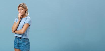 Studio shot of unconfident silly and insecure cute feminine girl with blond hair standing half-turned over blue background holding finger on lip while being shy ask question smiling hesitating photo