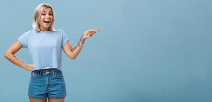 Studio shot of impressed and surprised entertained blonde female in summer outfit holding hand on waist pointing right and smiling joyfully being amazed and amused over blue background photo