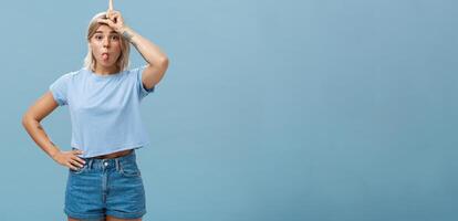 Studio shot of playful triumphing good-looking blonde female with tattoed arms showing letter L on forehead and sticking out tongue mocking over loser and celebrating own victory over blue wall photo