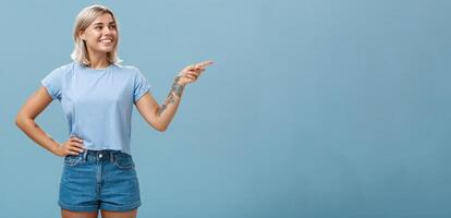 Bossy confident female manager with tattoo on arm holding hand on waist pointing and gazing right with pleased relaxed look giving directions to employees posing over blue background photo