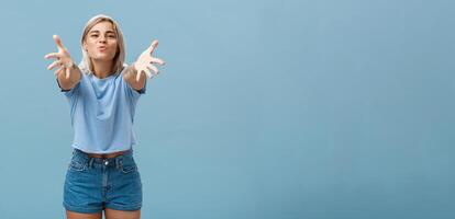 Come to mama. Portrait of friendly joyful and enthusiastic caucasian female student in trendy outfit pulling hands towads camera and folding lips to hug and give passionate kiss smiling over blue wall photo