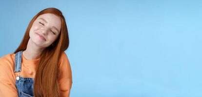 Dreamy kind silly redhead smiling happy girl straight long ginger hair daydreaming imagine romantic moment close eyes smiling delighted tilting head look joyful, standing blue background photo