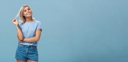 Studio shot of relaxed and chill popular attractive woman with blond hair and tattoos playing with strand tilting head and smiling as if listening boyfriend or flirting standing amused over blue wall photo