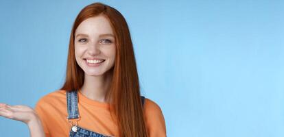 Proud good-looking confident redhead girl present awesome product hold object palm raise hand blank blue copy space smiling delighted recommend cool link, standing studio background helpful photo