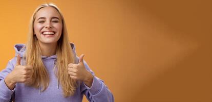 Close-up carefree delighted positive lucky young blond woman in hoodie close eyes joyfully celebrating good result show thumbs-up gesture agree approving awesome idea, orange background photo