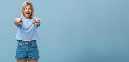 Happy enthusiastic stylish blonde female with tattoos pointing at camera as if picking or making hint smiling joyfully and winking from amazement and happiness posing over blue background photo