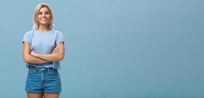 Ambitious good-looking young tanned female with blond hair standing in confident pose with hands crossed on chest smiling broadly standing in denim shorts over blue background photo