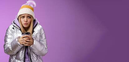 Trembling uncomfortable cute pouting young blond girl feel freezing cold winter snowy weather outside shaking low temperature hold take-away hot coffee cup warming up, purple background photo