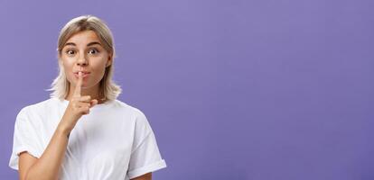 Charming creative and stylish beautiful tanned girl with fair hair shushing at camera with delighted thrilled look making up great idea wanting hide it and keep secret posing over purple wall photo