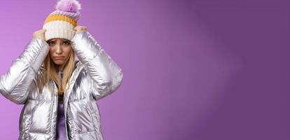 Upset gloomy insulted cute blond girl complaining offensive behaviour pulling hat forehead frowning sulking sad standing miserable pity purple background in warm stylish glittering silver jacket photo
