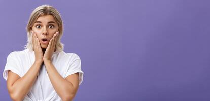 Waist-up shot of stunned impressed gorgeous tanned female in white t-shirt with blond hair gasping with opened mouth from surprise holding hands on cheeks from amazement and curiosity photo