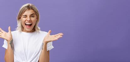 Waist-up shot of playful energized sociable attractive woman with blond hair, tanned skin and tattoos raising hands joyfully and spread aside winking from amusement smiling broadly over purple wall photo