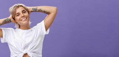 Close-up shot of good-looking stylish and relaxed blond woman with tattoos on arms holding hands behind head smiling with carefree pleased look enjoying weekends over purple background photo