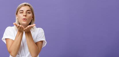 Blowing sweet kiss to all fans. Flirty tender and cute stylish european female with blond hair and tanned skin holding palms near mouth folding lips while sending mwah at camera over purple wall photo