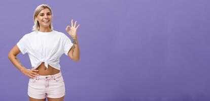 Excellent I like it. Portrait of satisfied good-looking happy girl with fair hair in white t-shirt and shorts showing ok or perfect gesture and smiling broadly holding hand on waist over purple wall photo