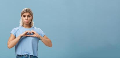 Heart being broken. Sad and gloomy heartbroken girl with blond hair tattoos on arms and tanned skin pursing lips whining and complaining making love sign over breast standing unhappy near blue wall photo