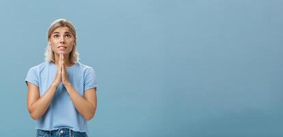 Studio shot of hopeful focused dreamy girl with attractive face and blonde hair holding hands in pray near body looking up hopefully with faith praying or making wish over blue background photo