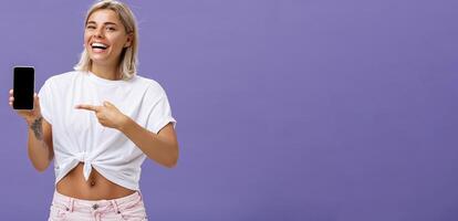 Satisfied happy beautiful tanned woman with blond hairstyle and tattoos holding smartphone showing device screen and pointing at gadget with index finger smiling at camera over purple wall photo