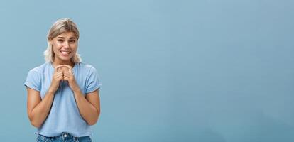 Silly cute and worried blonde female friend with short haircut steepling index fingers and smiling with guilty sorry smile frowning while waiting for answer begging for help or favor over blue wall photo