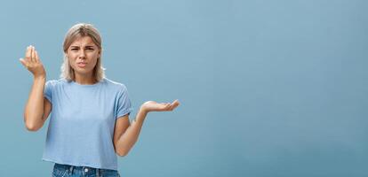 Wtf you saying. Portrait of pissed and confused stylish girlfriend with blond hair raising palm high and aside with perplexed expression being questioned hearing stupid nonsense over blue wall photo