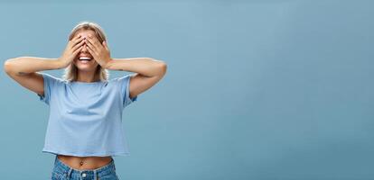 Hide if you can. Portrait of carefree and happy good-looking playful girl with blonde hair smiling broadly while counting ten and playing hide-and-seek with closed eyes over palms near blue wall photo