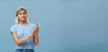 Woman cannot stand when mom shouts during phone call. Intense displeased young european blonde with tanned skin clenching teeth and frowning covering microphone of smartphone over blue wall photo