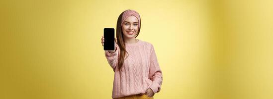 mejor teléfono inteligente aquí usted ir. encantador saliente joven bonito mujer en de punto suéter, venda extensión mano con teléfono demostración artilugio pantalla sonriente recomendando Teléfono móvil terminado amarillo pared foto