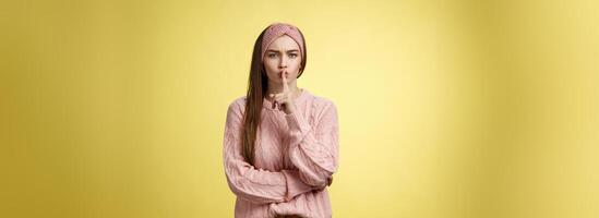 Keep mouth shut. Serious-looking bossy attractive young 20s woman in sweater, headband shushing making shhh gesture holding index finger on lips, gossiping, spread rumors over yellow background photo