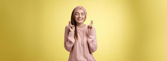 Girl wishing wellness wanting dream come true, cross fingers dreamy, close eyes waiting miracle, anticipating good news, posing excited and joyful against yellow background in knitted warm sweater photo