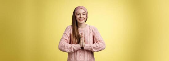 Por favor hacer yo favor. encantador joven hembra estudiante vistiendo de punto suéter, venda prensado palmas juntos en orar mendicidad, preguntando para ayuda sonriente agradecido, agradecido para esfuerzo terminado amarillo pared foto