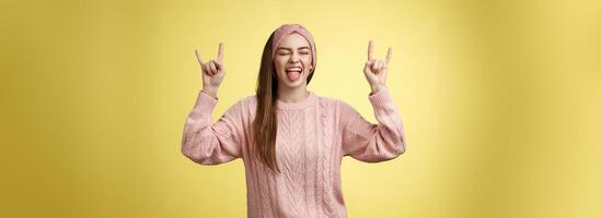 Cute heavy metal lover showing rock roll symbol sticking tongue amused and happy fooling around listening favourite music posing excited and pleased against yellow background in knitted outfit photo