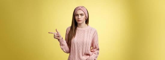 Pissed puzzled young attractive arrogant moody girlfriend in sweater, headband looking irritated, intense frowning pointing finger at copy space, directing person sits on her place, annoyed photo