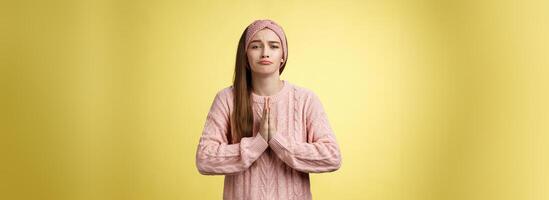 Deliberately sad girl in sweater pressing palms together in pray pouting praying for help, asking favor, begging friend lend cute dress for date, smiling silly, want something over yellow background photo