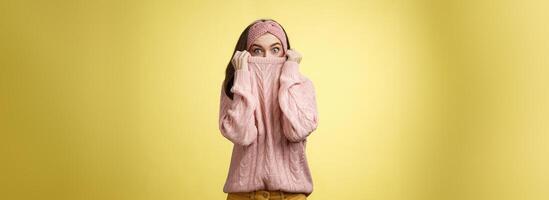 Girl hiding in collar wearing knitted sweater over nose popping eyes, staring bugged eyes at camera, hiding face reacting amazed and shocked, feeling scared, standing in stupor over yellow background photo
