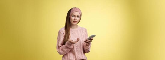 Puzzled confused upset questioned young cute girl in headband knitted sweater frowning clueless raising hand in dismay holding smartphone, cannot understand what happened cellphone over yellow wall photo