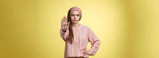 Stop it. Serious-looking confident focused young cute girl extending arm hold, tresspass gesture, frowning displeased, standing bossy denying, forbidding pass, unwilling let-through over yellow wall photo