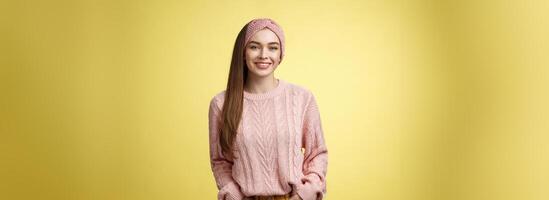 Charming friendly young girl in knitted sweater and headband holding hands in pockets, smiling, looking positive motivated achieving goals having great day, posing positive against yellow background photo