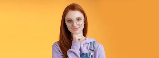 Confident happy relaxed european redhead skillful young female entrepreneur glasses establish startup smiling delighted look pleased result touch chin thoughtful smart, orange background photo