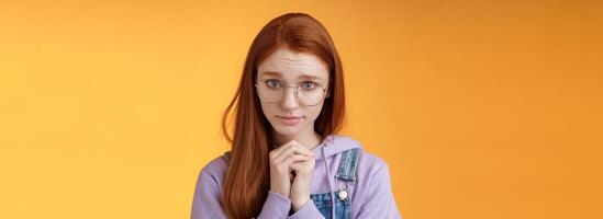 Silly guilty young shy redhead girlfriend asking forgiveness supplicating lower head look from under forehead frowning begging apology favour standing insecure sad pleading help, orange background photo