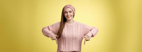 oferta joven 20s europeo niña en suéter sonriente en general finalmente quitarse tirantes señalando abajo promoviendo hábil dentista profesional. hembra estudiante en pie contento positivo terminado amarillo pared foto