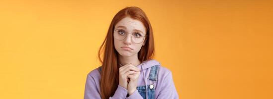 Pretty please. Clingy young sad sulking redhead girl sibling wearing glasses pouting silly press palms begging gesture pleading wanna receive help promise be good ask favour, orange background photo