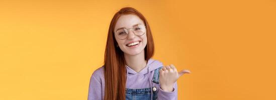 Cute helpful friendly-looking joyful european redhead woman show thumb left smiling delighted laughing pointing where find awesome store telling about interesting new promo offer, orange background photo