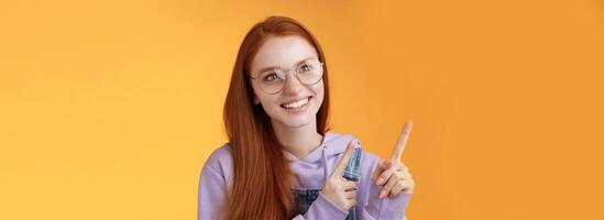 Carefree charismatic happy silly young cute redhead girl freckles blue eyes wearing glasses enjoy contemplating entertaining exhibition pointing looking left indicating, orange background photo