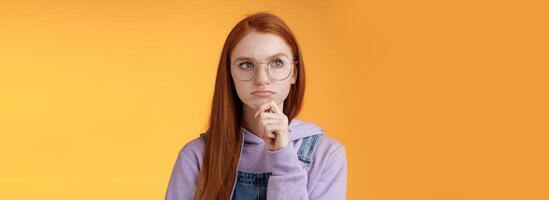 perplejo de aspecto serio inteligente joven pelirrojo niña confuso pensando profundo atención problema toque barbilla ceñudo Mira Superior izquierda esquina considerado, reflexionando suspicaz situación, naranja antecedentes foto