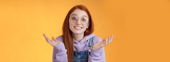 Well no big deal. Attractive silly redhead hipster girl wearing glasses smiling awkward unaware spread hands sideways shrugging confused grinning questioned uncertain what tell, orange background photo